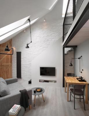 Pokój dzienny w poddaszowym małym mieszkaniu z szarą sofą elementami z drewna czarnymi lampami i antresolą z czarną metalową balustradą