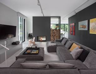 Duże miękkie szare sofy w pokoju dziennym ciemnoszara ściana i minimalistyczny kominek w domu z dużymi oknami