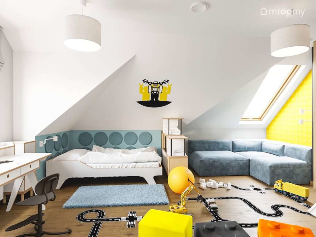 Łóżko z białej sklejki z miękkimi panelami na ścianie naklejką lego i niebieskim dywanem w poddaszowym pokoju chłopca