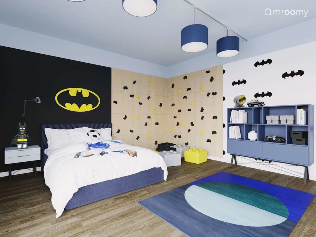 Ścianka wspinaczkowa obok łóżka nad którym jest żółta naklejka batman w pokoju chłopca w którym są niebieskie meble na cienkich nóżkach