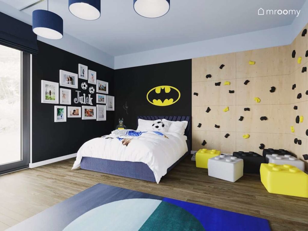 Czarna ściana z naklejką Batmana duże łóżko i ścianka wspinaczkowa z pufami lego w pokoju wczesnoszkolnego chłopca
