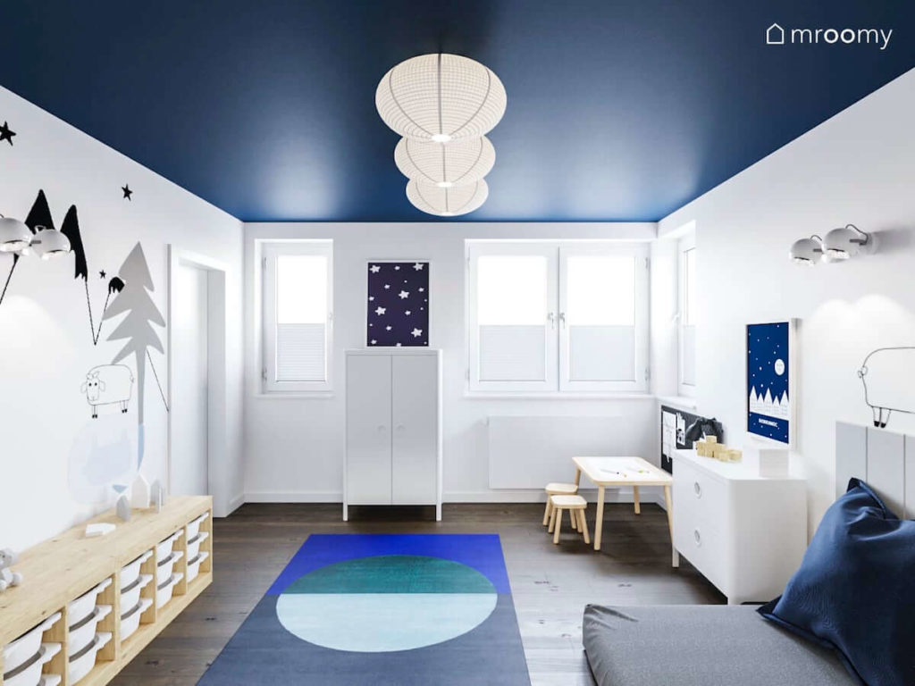 Stolik do rysowania regał na zabawki niebieski dywan biała szafa i miejsce do spania w minimalistycznym pokoju dla małego chłopca