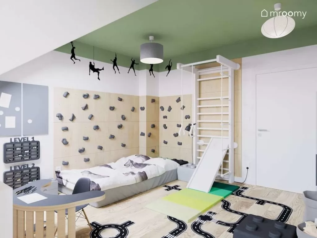 Ścianka wspinaczkowa przy łóżku w pokoju małego chłopca w którym jest również biała drabinka gimnastyczna i zielony sufit