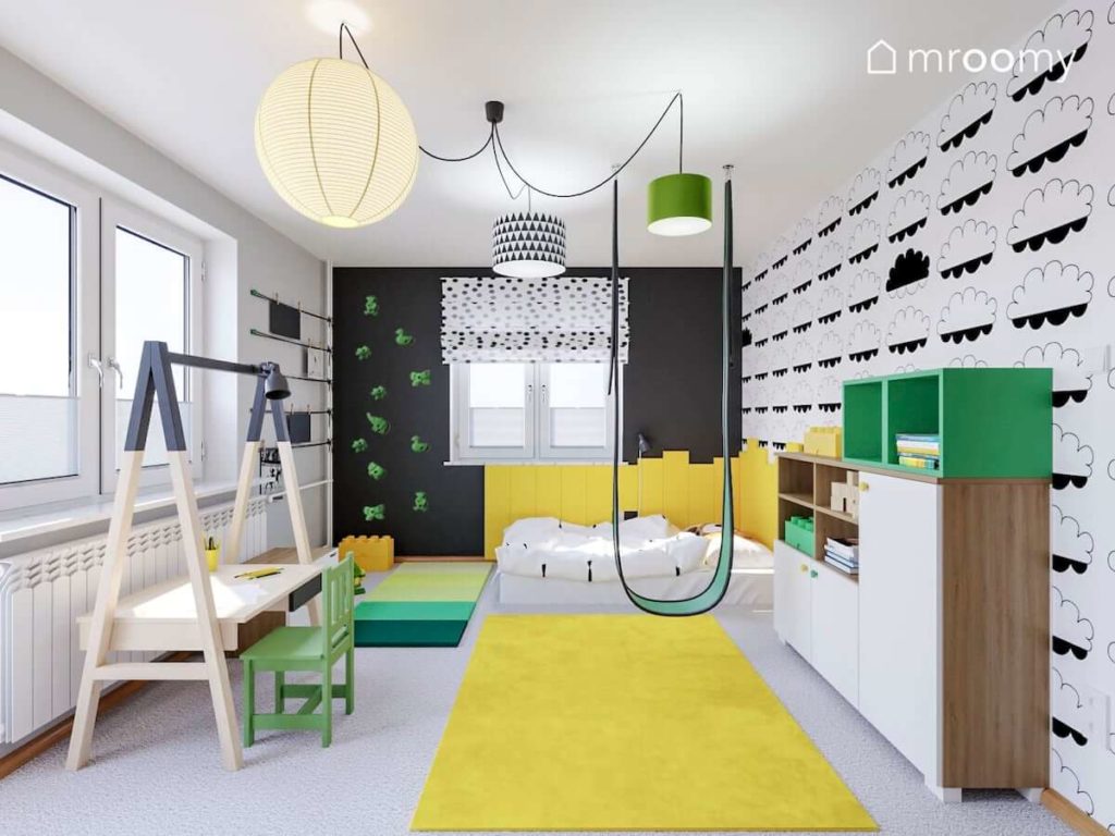 Czarna ściana z zielonymi chwytami wspinaczkowymi  biurko rosnące z dzieckiem i żółte akcenty w pokoju kilkuletniego chłopca