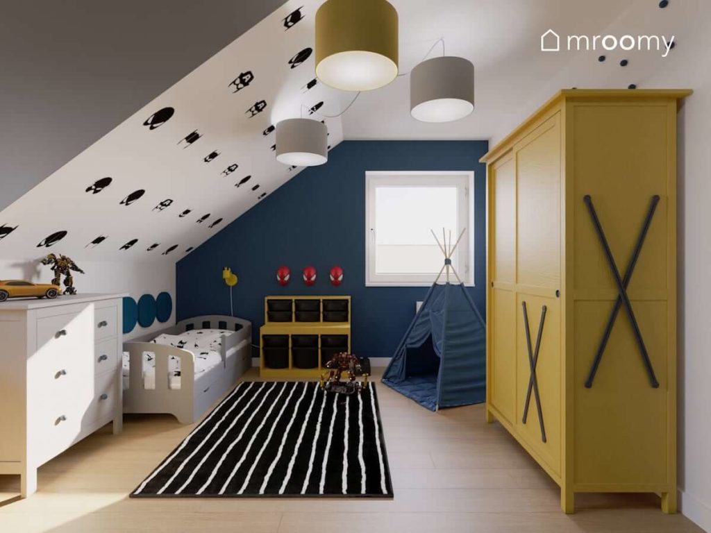 Żółta szafa niebieska ściana i lamy spiderman w pokoju chłopca ze skosami i szarym łóżkiem obok czarnego dywanu