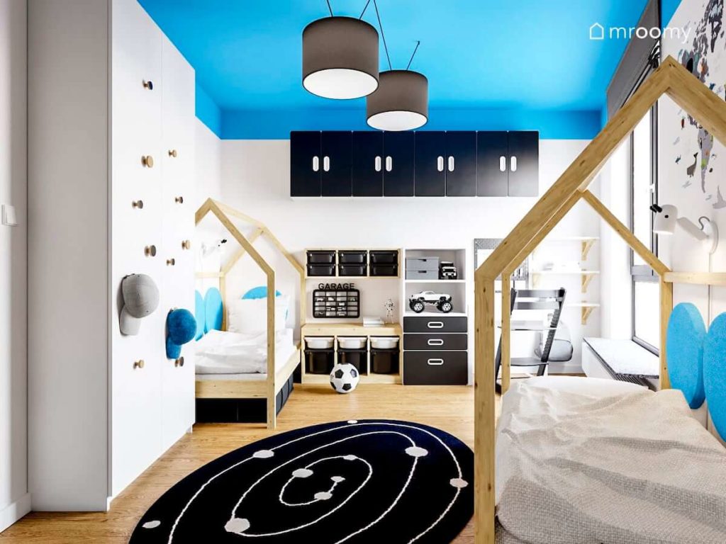 Białe meble z czarnymi elementami dwa łóżka domki i dywan z motywem kosmosu w pokoju dwóch małych chłopców z niebieskim sufitem