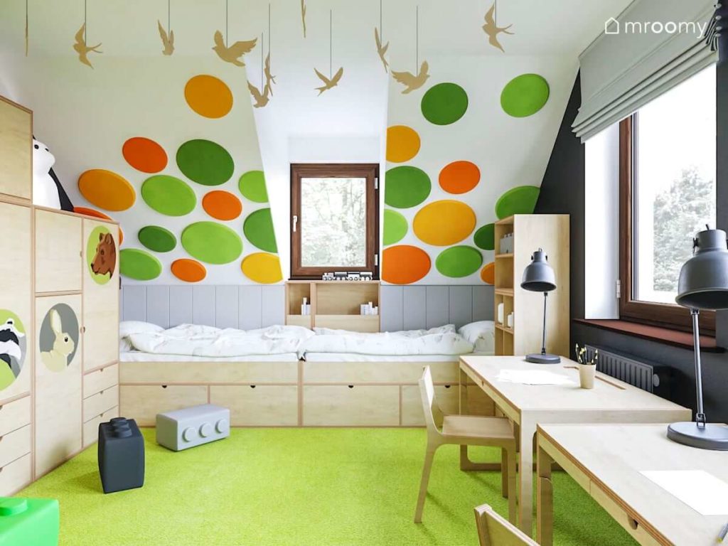 Kolorowe panele ścienne nad dwoma łóżkami obok szafy i biurek ze sklejki w pokoju ze skosami dwóch małych chłopców
