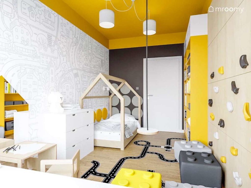 Łóżko domek przy ścianie z czarno-białą tapetą w miasto i ulice w pokoju małej dziewczynki  w którym są również ścianka wspinaczkowa i żółto-białe meble