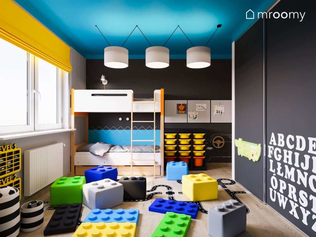 PIętrowe łóżko w czarno-niebiesko-żółtym pokoju dwójki małych dzieci z miękkimi pufami w kształcie lego i niebieskim sufitem