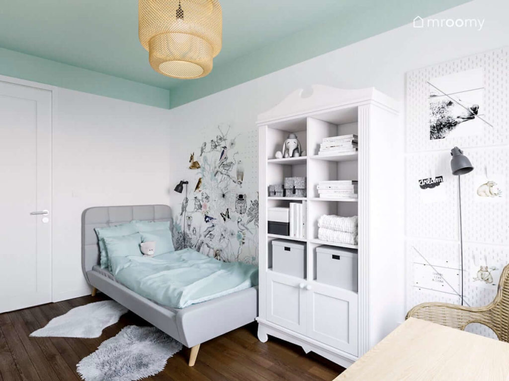 Szare łóżko graficzna tapeta z drzewem i biały klasyczny regał z miętowymi gałkami w romantycznym pokoju dziewczynki