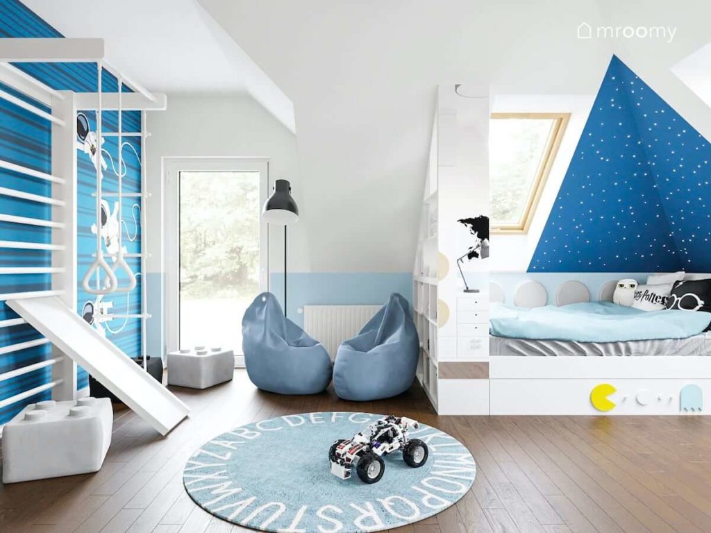 Biała drabinka gimnastyczna przy niebieskiej ścianie worki sako okrągły dywan i łóżko przy ścianie ze skosem w pokoju chłopca