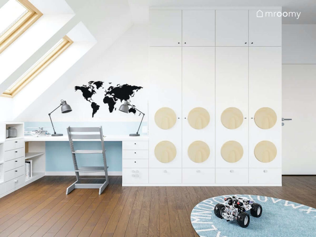 Biurko przy ścianie ze skosem w pokoju chłopca nad którym jest naklejka z mapą świata obok biała szafa z okrągłymi panelami