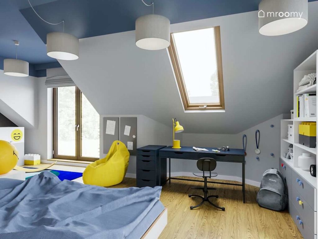 Granatowe biurko pod oknem połaciowych obok żółty worek sako organizery magnetyczne na ścianie i niebieski sufit w pokoju wczesnoszkolnego chłopca