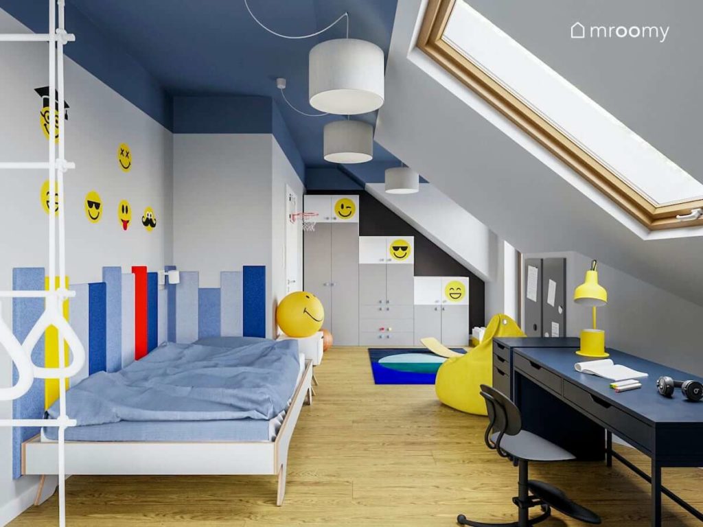 Łóżko granatowe biurko i szafa pod skosem w pokoju chłopca z niebieskim sufitem i żółtymi dodatkami