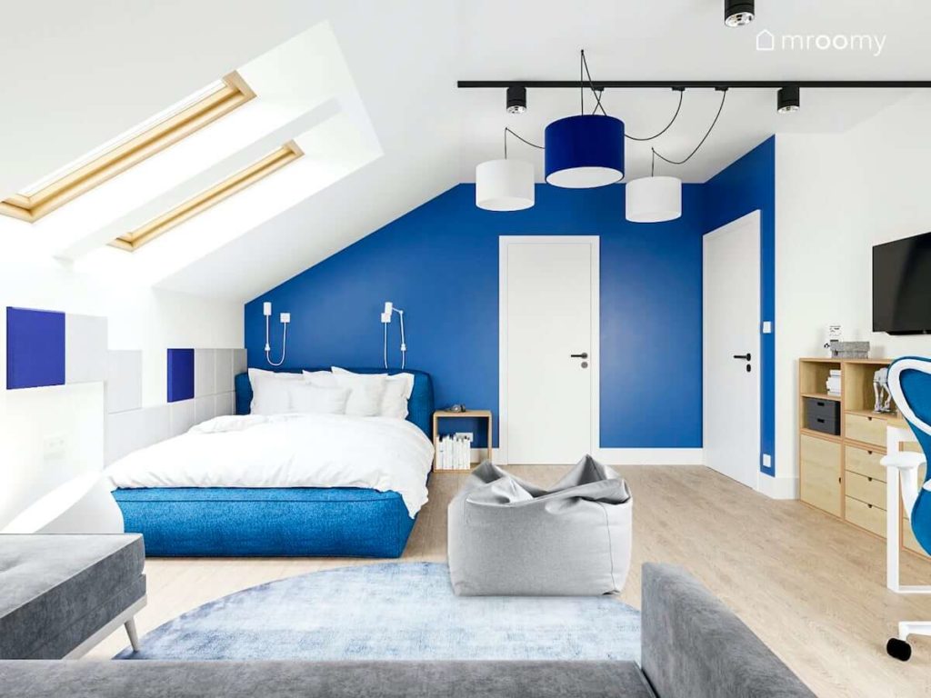 Miękkie duże tapicerowane niebieskie łóżko niebieska ściana i fotel sako w dużym poddaszowym pokoju nastolatka