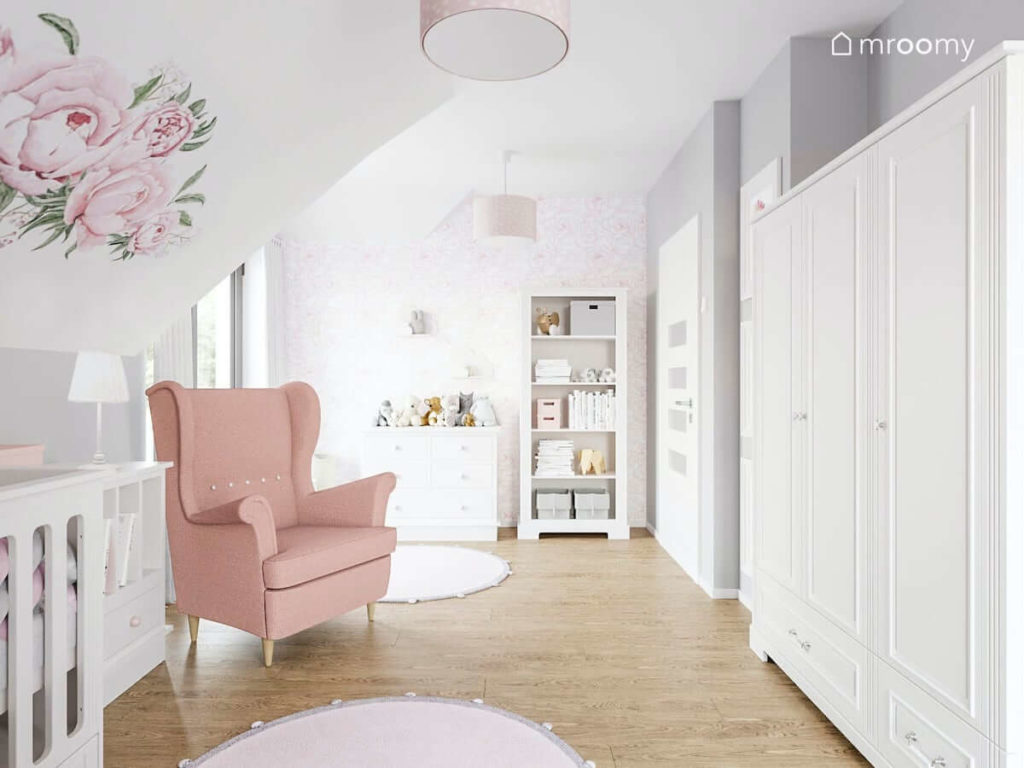 Biała szafa regal i komoda obok różowy fotel dla mamy w pokoju malutkiej dziewczynki na poddaszu z różowymi okrągłymi dywanami