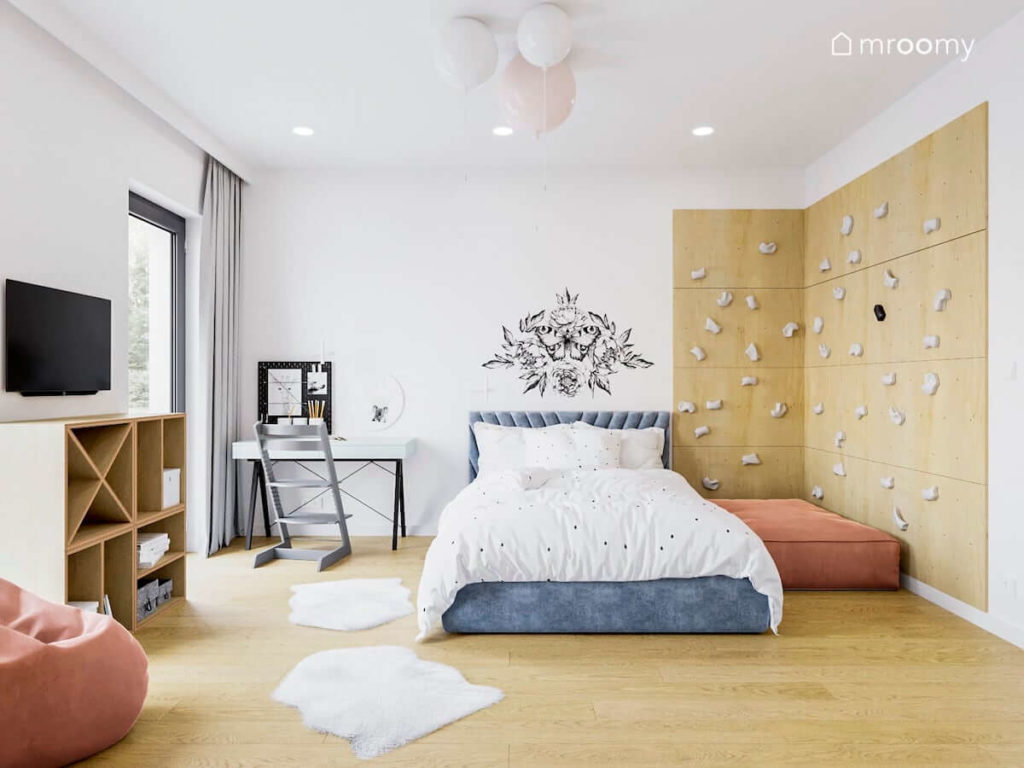 Ścianka wspinaczkowa obok tapicerowanego łóżka i białego biurka w pokoju dla dziewczynki