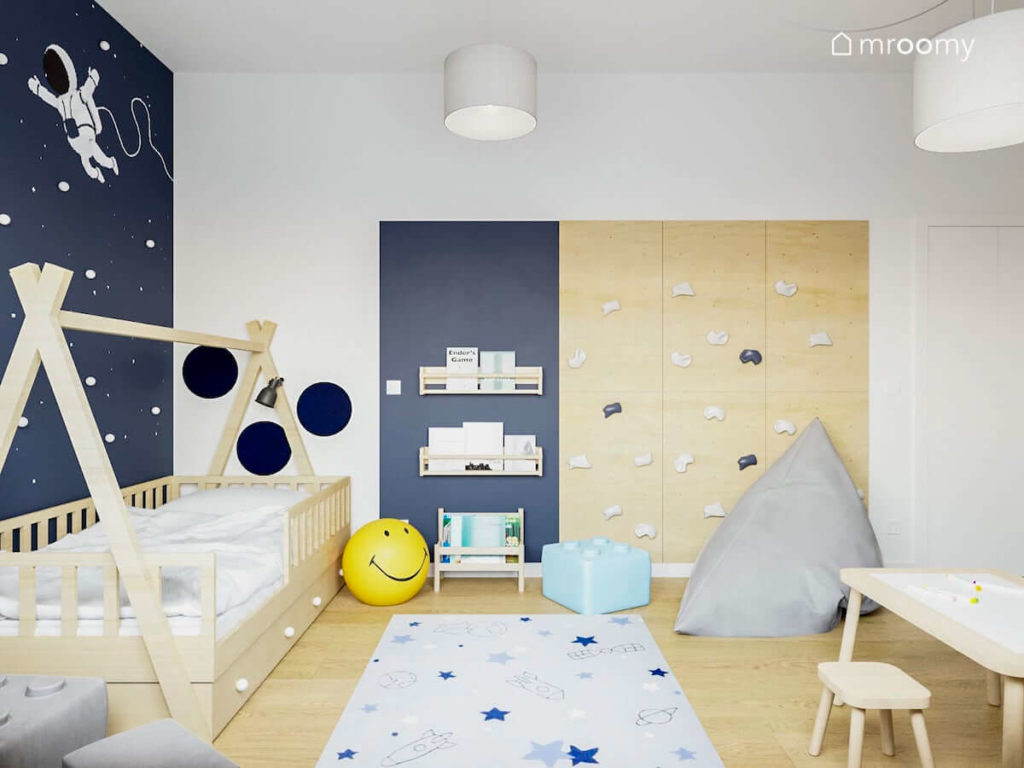 Ścianka wspinaczkowa obok niebieskiej ściany i łóżka domku worka sako i lampy w uśmiechniętą buźkę w pokoju małego chłopca