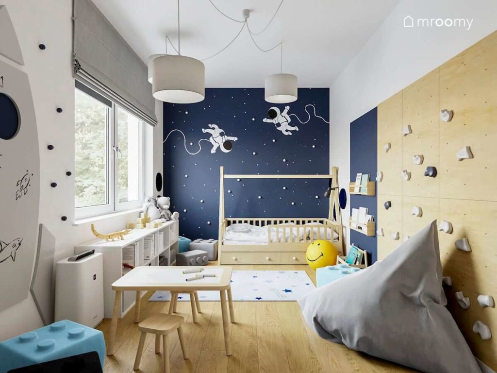 Niebieska ściana z gwiazdami i astronautami w pokoju chłopczyka z łóżkiem tipi stoliczkiem i ścianką wspinaczkową
