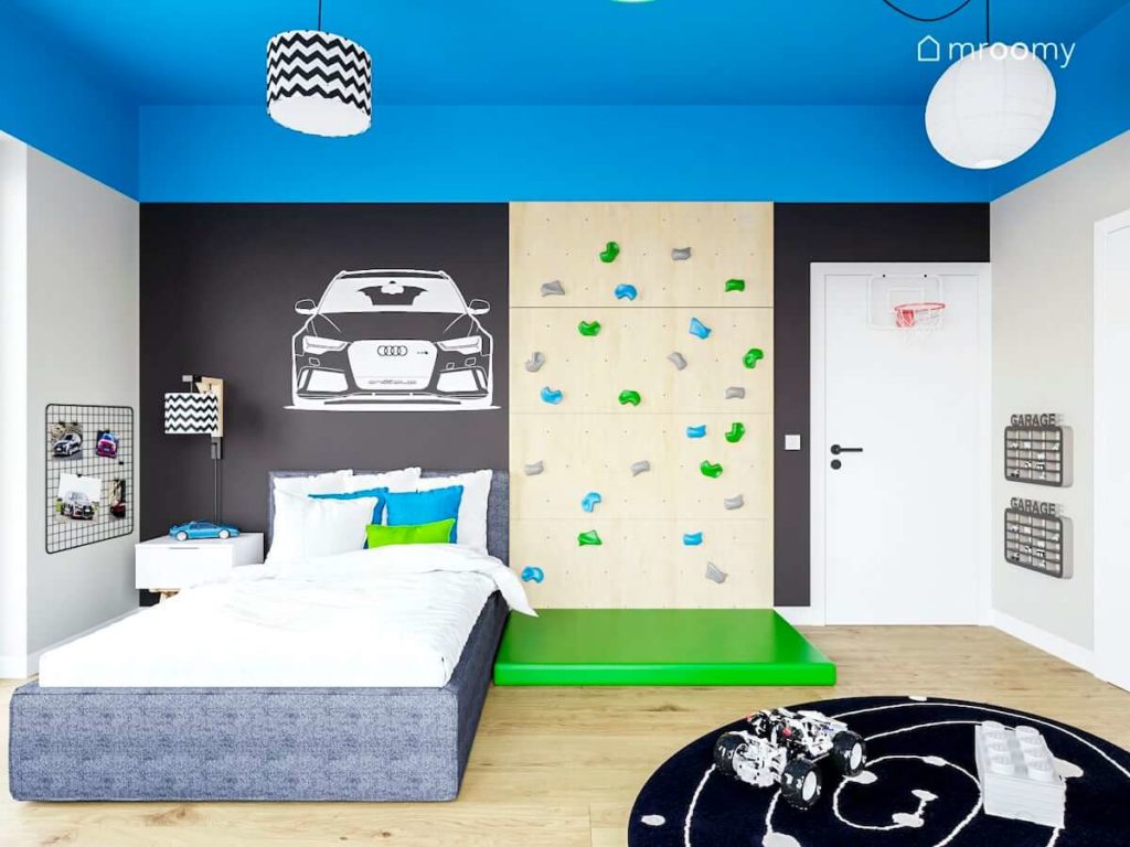 Ciemne ściany ze ścianką wspinaczkową dużym łóżkiem kolorowym sufitem i kolorowe abażury w pokoju chłopca