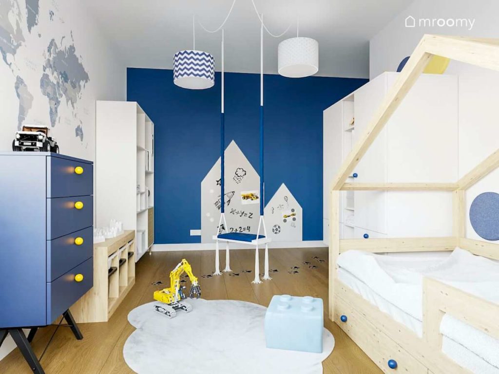 Łóżko domek białe meble i niebieska ściana z tablicą suchościeralną przy huśtawce w pokoju małego chłopca