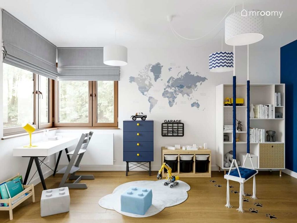Granatowa komoda obok białego regału przy ścianie z tapetą z mapą świata narożne okno i huśtawka w pokoju dziecka małego chłopca