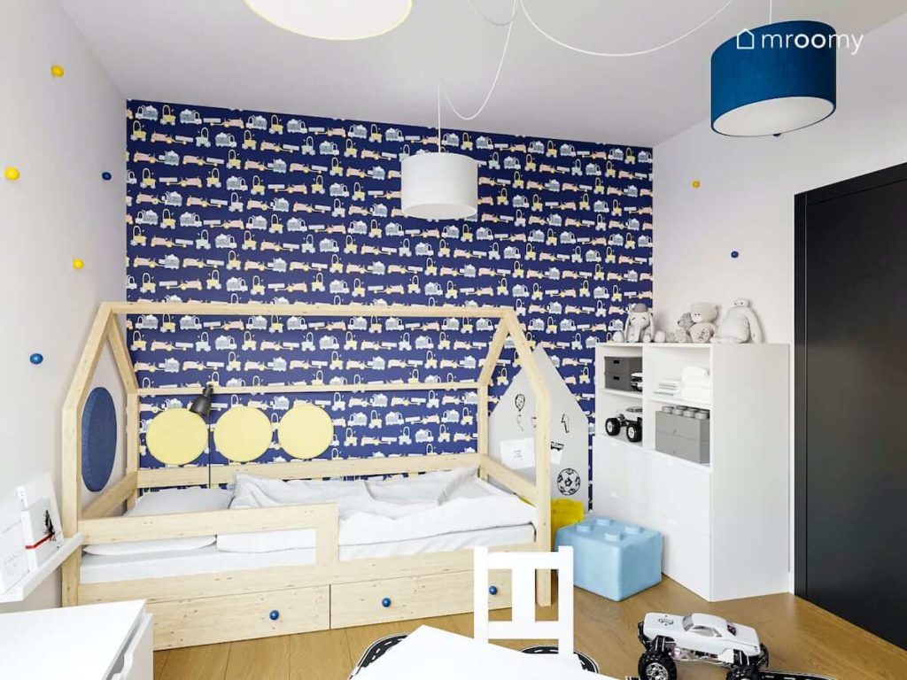 Ściana w granatową tapetę z autkami obok łóżko domek i biały regał w pokoju dziecka przedszkolaka