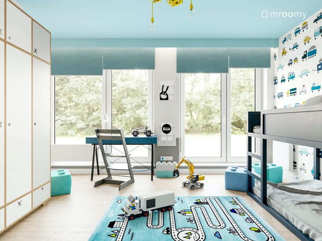 Pokój małego chłopca z dużym oknem balkonowym przy którym stoi niebieskie biurko i pufy lego na podłodze