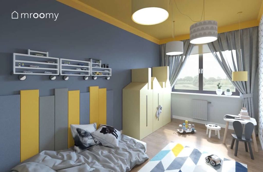 Żółta szafa w kształcie kamieniczki miękkie panele ścienne obok łóżka i kolorowy dywan w romby w pokoju dla małego chłopca