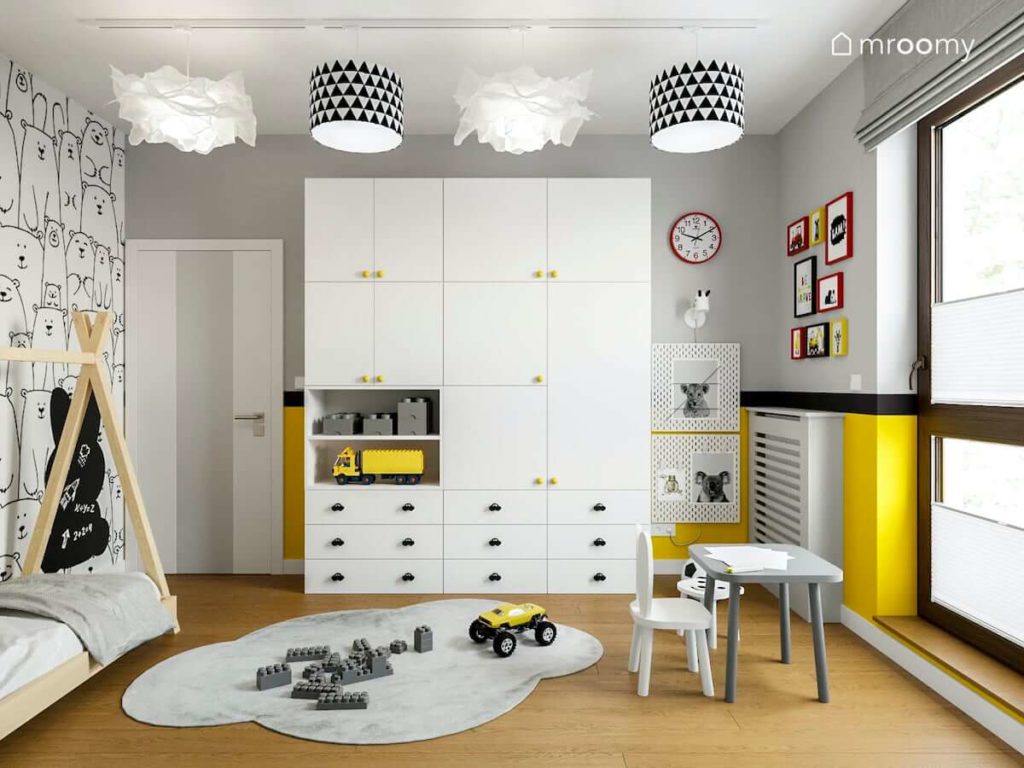 Biała szafa na tle białej i żółtej ściany szary dywan chmurka stolik z krzesełkiem i abażury w czarno-białe trójkąty w pokoju małego chłopca