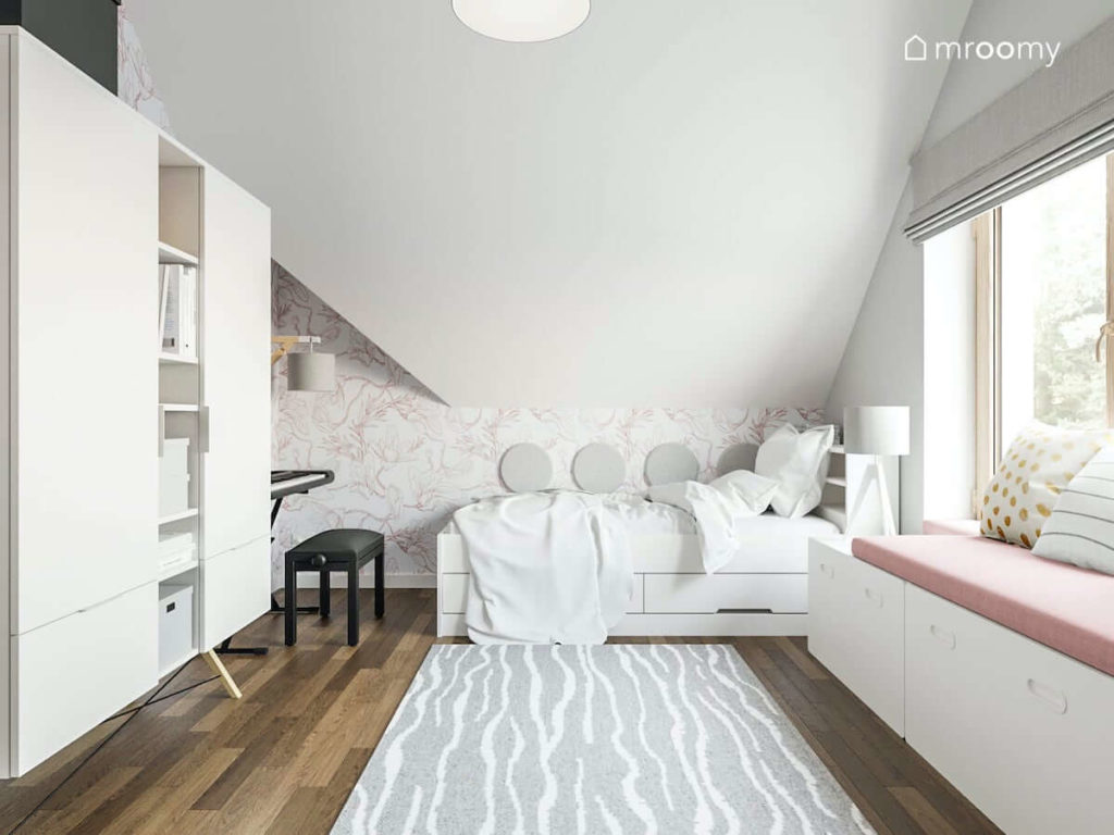 Ściana przy białym łóżku z miękkimi panelami ściennymi biała szafa na drewnianych nóżkach i szary dywan w pokoju nastolatki