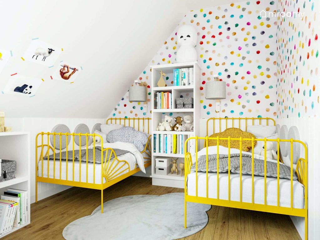 Dwa żółte łóżka metalowe pod skosem na tle tapety w kolorowe kropki i dywanik chmurka w pokoju dziewczynek sióstr