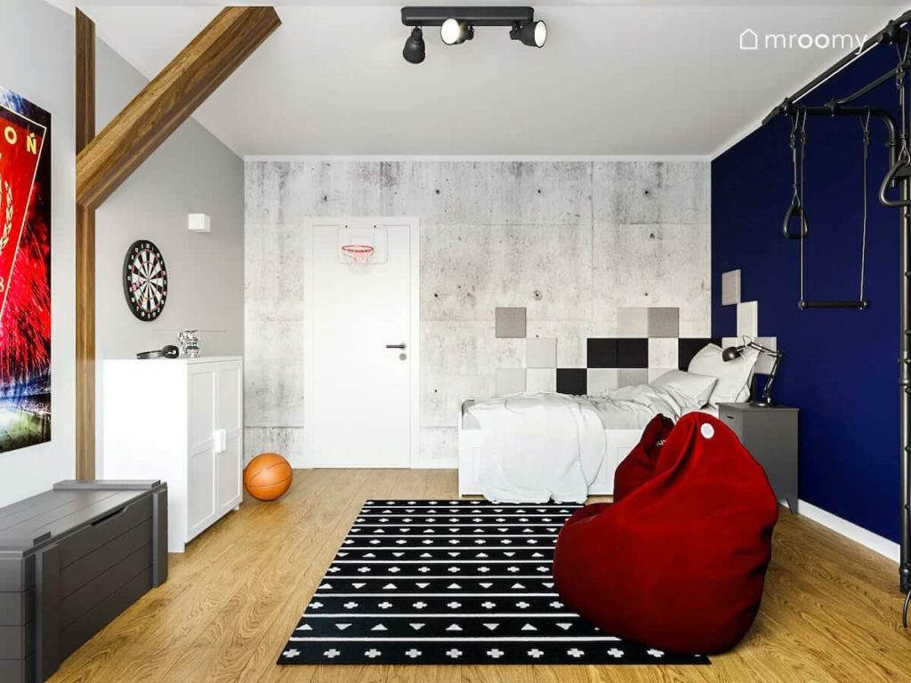 Pokoj nastolatka z tapeta imitujaca betonowe plyty bialymi meblami i granatowa sciana Bordowy worek sako i czarno bialy dywan w pokoju chlopca