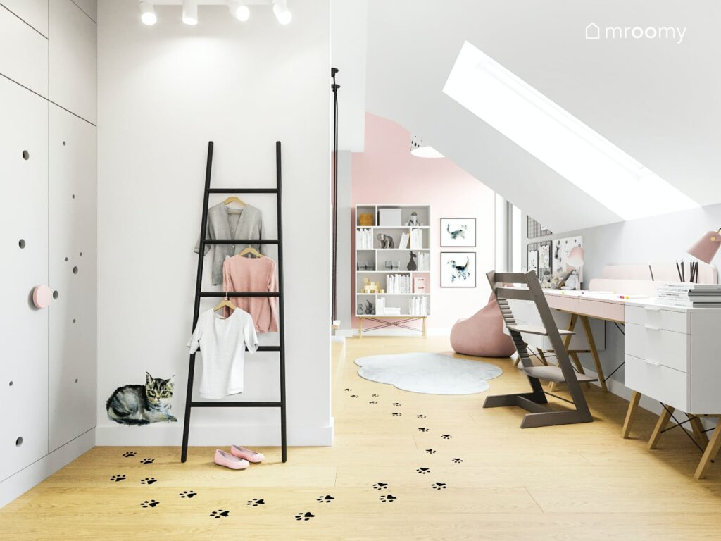 Przestronny pokój dla dziewczynki utrzymany w kolorach jasnoróżowym i białym w nim pojemne białe szafy oraz regały pod oknem biurko na podłodze naklejki w kształcie łapek przy ścianie drabinka wieszak i naklejka ścienna kot