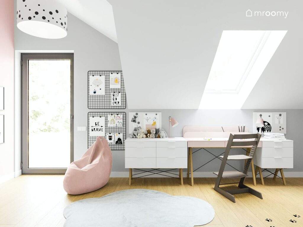 Przestronny jasny pokój dla dziewczynki ze ścianami w kolorze białym i jasnoróżowym pod oknem dachowym długie biurko na podłodze szary dywan oraz różowa wygodna pufa obok biurka organizery a na suficie duża lampa wisząca