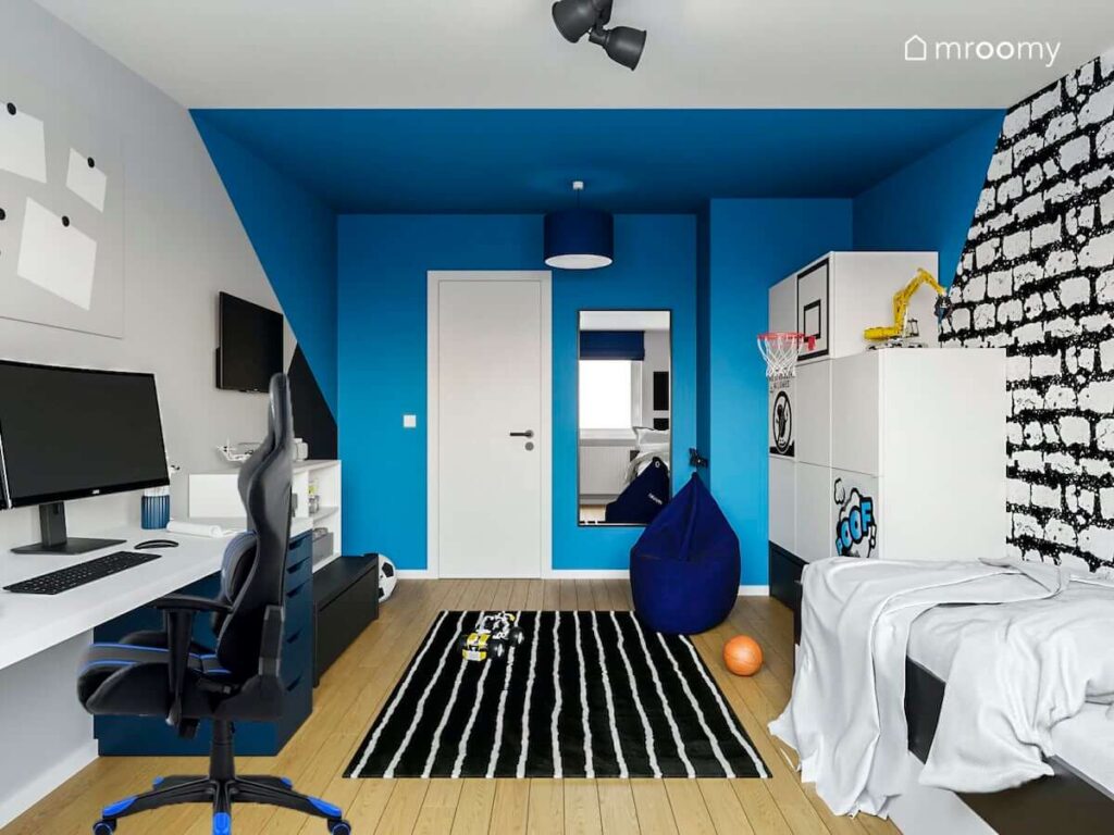 Biało niebieski pokój nastolatka z dużym biurkiem łóżkiem i białą szafą z metalowymi nakładkami na frontach a na podłodze czarno biały dywan i pufa sako