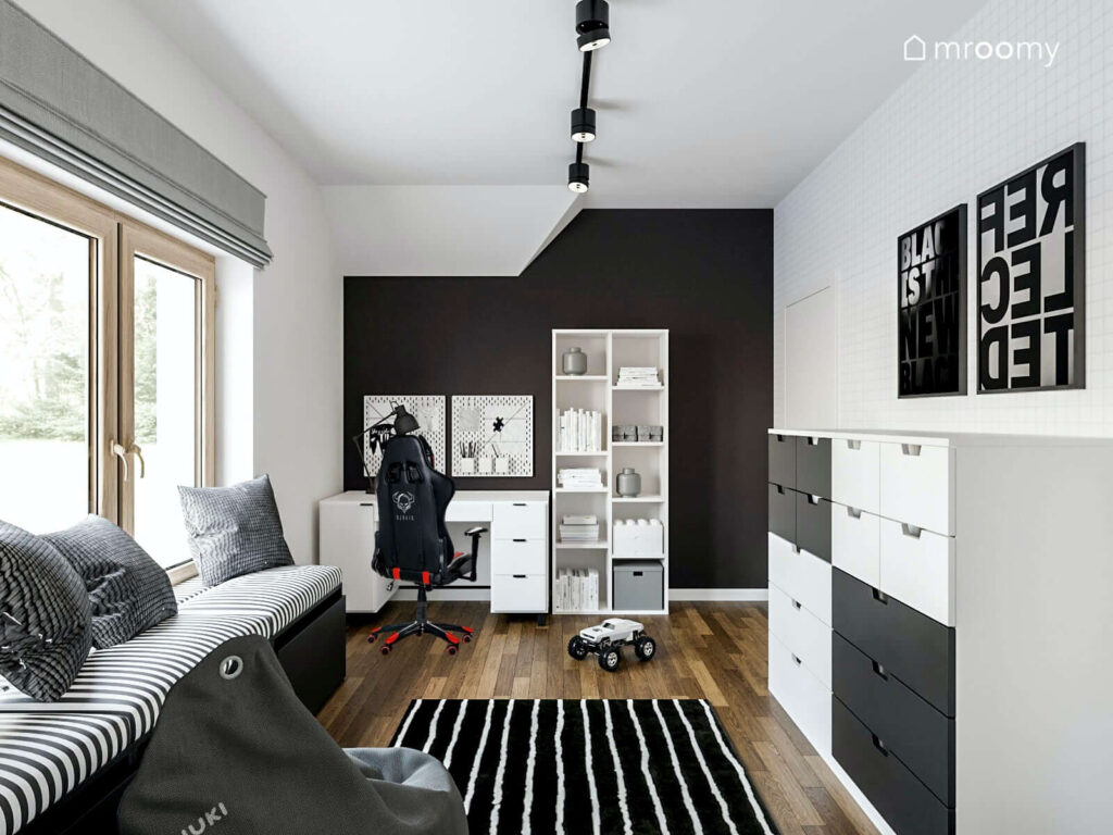 Pokój dla nastolatka utrzymany w tonacji czarno białej z tapetą w zeszytową kratkę a w nim komoda regał oraz biurko z szufladami a także tapicerowane siedzisko dywan w paski i pufa sako