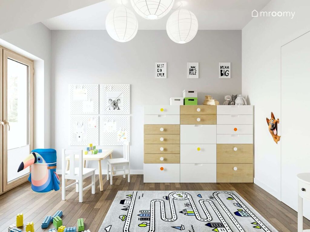 Jasny pokój dla małego chłopca z komodą z wieloma szufladami stolikiem dziecięcym oraz dywanem we wzór z drogą na ścianie praktyczne organizery a na suficie lampy w papierowych kloszach