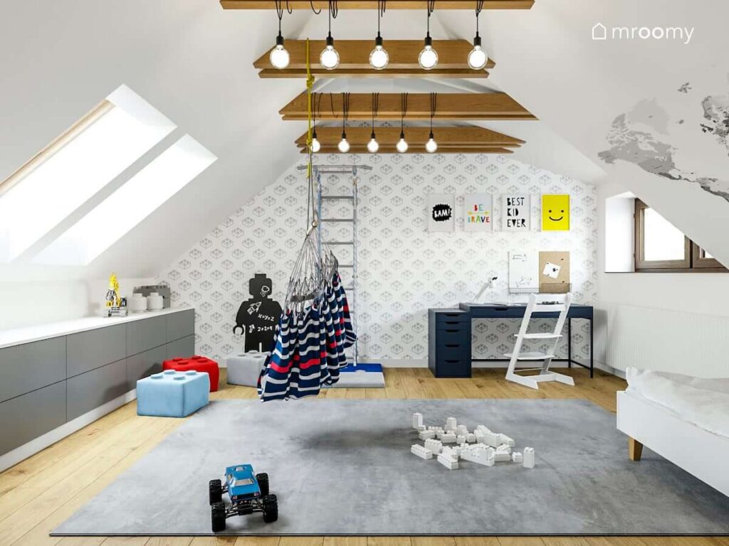 Pokój na poddaszu dla chłopca a w nim duży szary dywan biurko drabinka gimnastyczna i pojemne komody na ścianie tapeta w klocki Lego oraz ozdobne plakaty a na suficie lampy zawieszone na belkach sufitowych