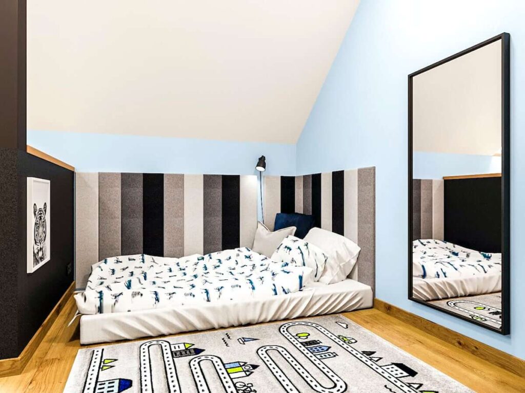Niskie łóżko uzupełnione szarymi miękkimi panelami ściennymi oraz lustro i dywan z drogą w strefie odpoczynku w pokoju dla chłopca