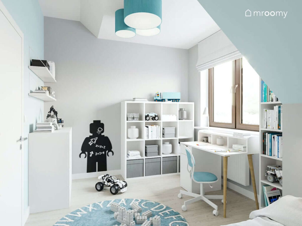 Biało niebieski pokój dla chłopca z pojemnymi regałami oraz komodą i funkcjonalnym biurkiem a także magnetyczną tablicą kredową w kształcie ludzika Lego