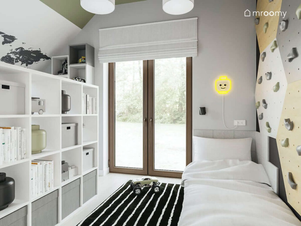 Pokój dla chłopca fana lego ze ścianką wspinaczkową nad łóżkiem i lampką w formie ledonu w kształcie główki ludzika lego z oknem z roletą rzymską