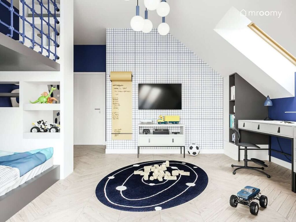 Ściana ozdobiona tapetą w kratkę łóżko na antresoli szafka RTV oraz biurko i szafki w biało granatowym pokoju dla chłopca
