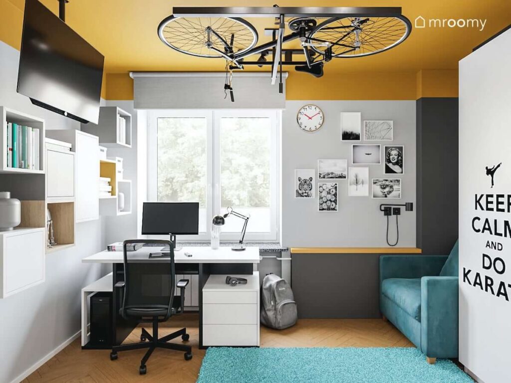 Pokój dla nastolatka z kolorowymi szafkami biurkiem rozkładanym fotelem oraz uchwytem na rower na pomarańczowym suficie i obrazkami w ramkach na ścianie