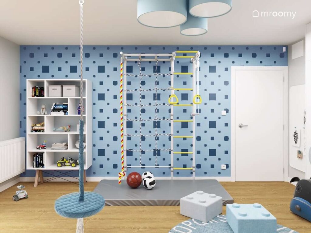 Kolorowa drabinka gimnastyczna z materacem oraz duży regał i zawieszona u sufitu huśtawka a na ścianie niebieska tapeta w kwadraty w pokoju kilkuletniego chłopca