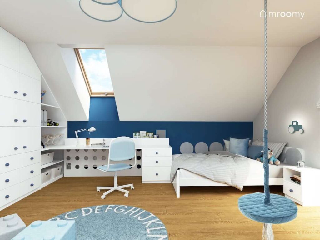 Biało niebieski pokój dla chłopca na poddaszu z pojemnymi regałami biurkiem łóżkiem oraz drewnianą huśtawką a na podłodze dywan z alfabetem