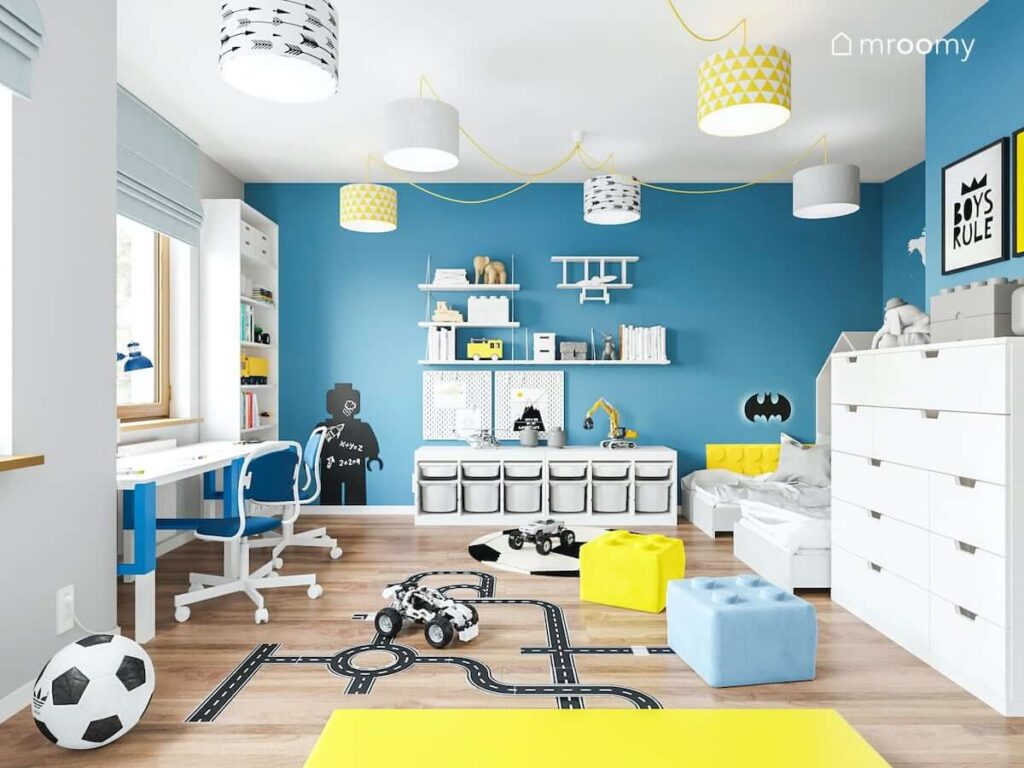 Biało żółto niebieski pokój dla chłopców a w nim długi regał z pojemnikami naklejka jezdnia a na ścianie kilka praktycznych półek i tablica kredowa w kształcie ludzika Lego