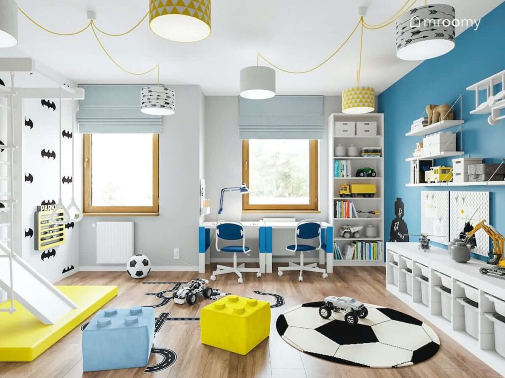 Kolorowy pokój dla dwóch chłopców z małymi biurkami wysokim regałem pufkami w kształcie klocków oraz kilkoma lampami sufitowymi