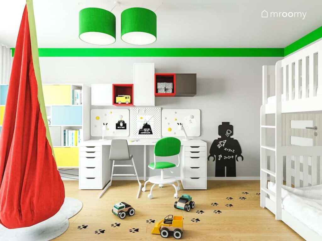 Strefa nauki z białym biurkiem organizerami kolorowymi półkami oraz tablicą kredową w kształcie ludzika Lego oraz zielona lampa sufitowa w pokoju dla dwóch chłopców