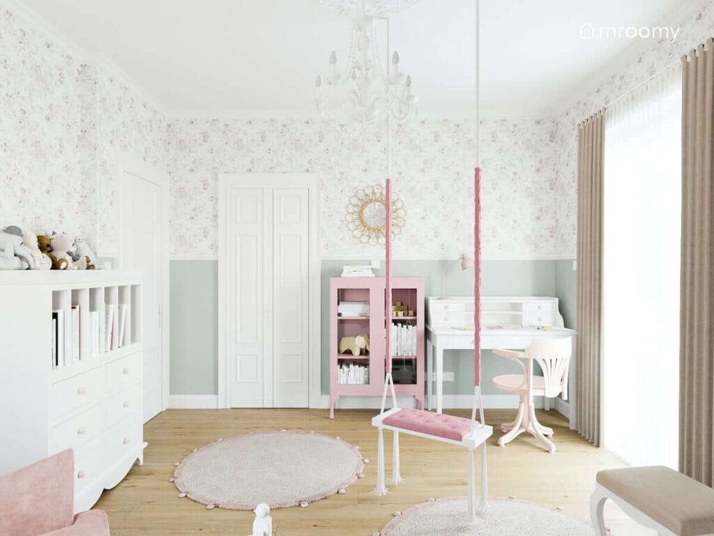 Jasny pokój dla kilkuletniej dziewczynki ze ścianami pokrytymi tapetą w kwiaty i szarą farbą zaś meble i dodatki w kolorach białym i różowym na środku pokoju dywany i wisząca huśtawka